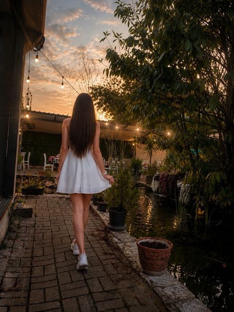 Viki Angels, gün batımında, üzerinde ışık dizileri asılı tuğla döşeli bir bahçe yolunda yürüyen beyaz bir elbiseyle