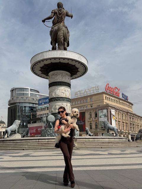 Viki Angels, Üsküp’te Makedonya Meydanı’nda, atlı heykelin altında, kahverengiler içinde küçük beyaz bir köpek tutuyor