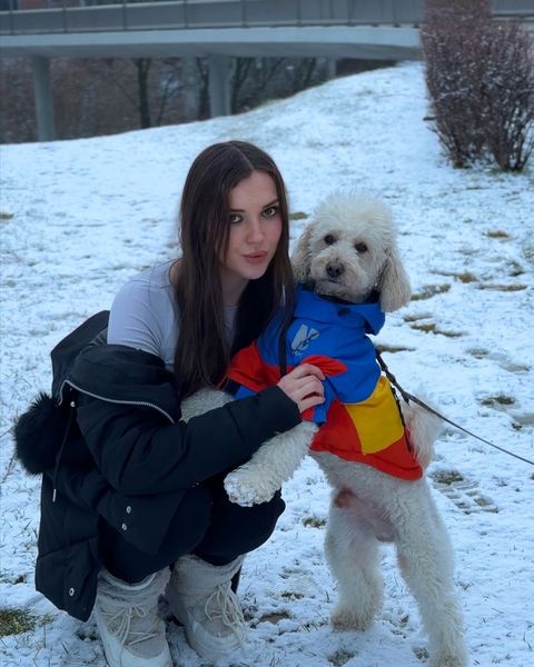 Viki Angels, karda çömelmiş, siyah şişme bir mont içinde küçük beyaz bir köpeğe sarılıyor
